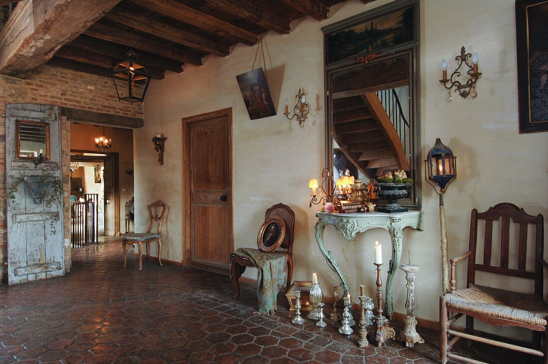 Rustikaler Hausflur eines französischen Landgutes mit verschiedenen antiken Holzstühlen und Spiegel über einem Rokoko-Konsolentisch