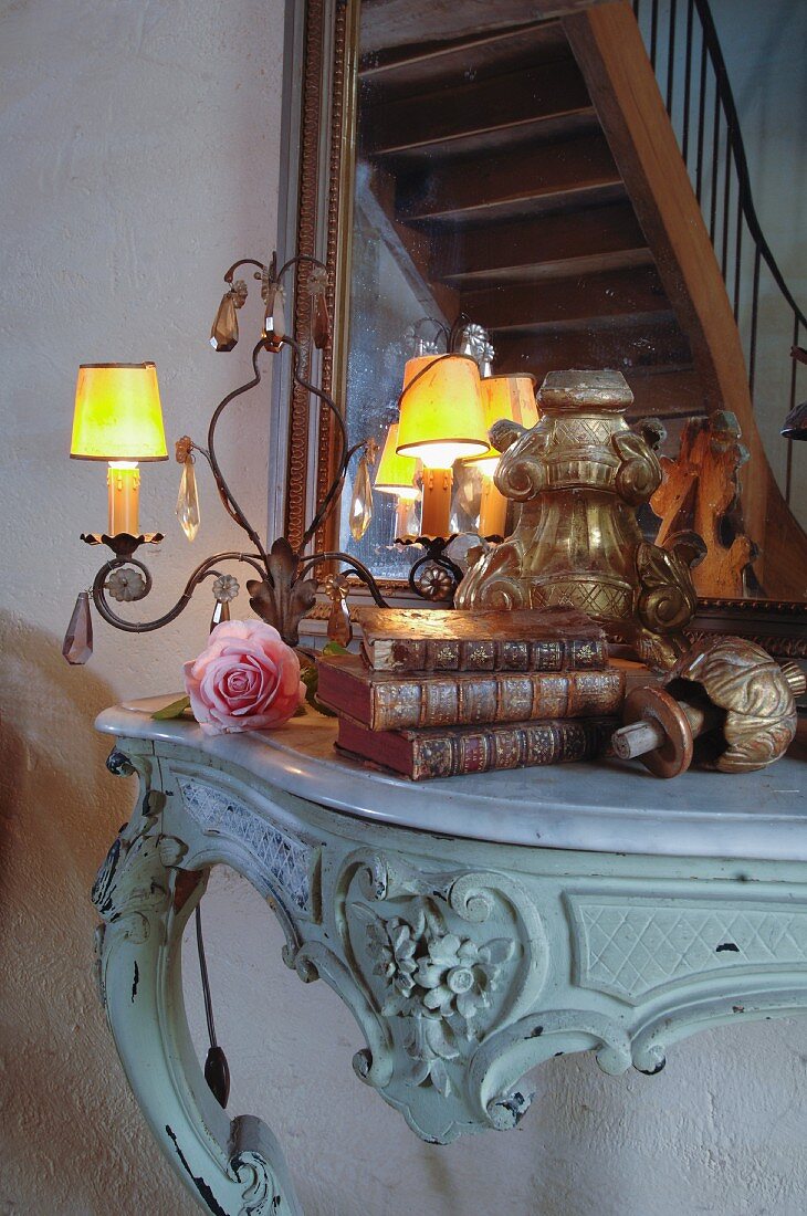 Sammlung antiker Flohmarktstücke und brennende Lämpchen auf Rokoko-Konsolentisch; Treppenreflektion auf Spiegel im Hintergrund