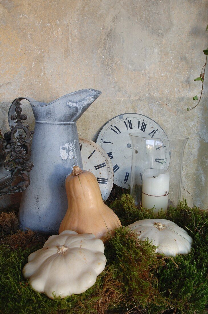 Antiker Wasserkrug, Ziffernblätter alter Uhren und Kürbisse auf Moosbett als romantische Landhausdekoration