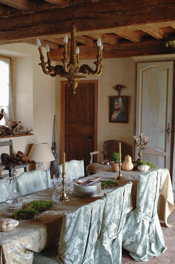 Festliche Dekoration auf der mit edlem Stoff drapierten, langen Tafel und romantische Hussenstühle in rustikaler Wohnküche