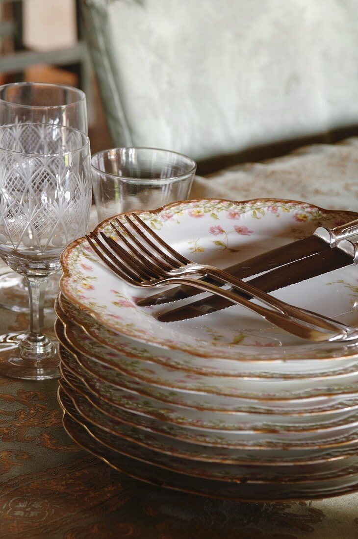 Stack of romantic floral plates and cutlery; old wine glasses in background