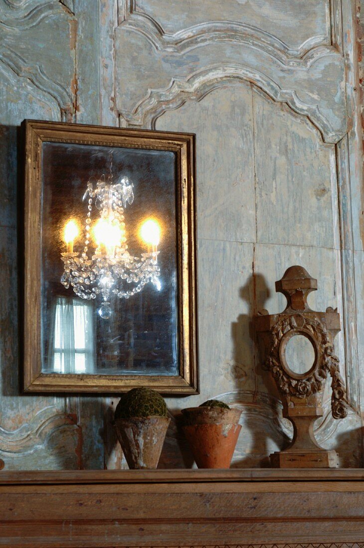 Gilt-framed mirror on faded wood-panelled wall and terracotta pots next to wooden sculpture on table top