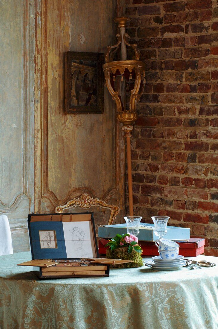 Frühstücksgeschirr neben aufklappbarem Kästchen mit Malutensilien auf Tisch und Prozessionsstab rustikaler Zimmerecke