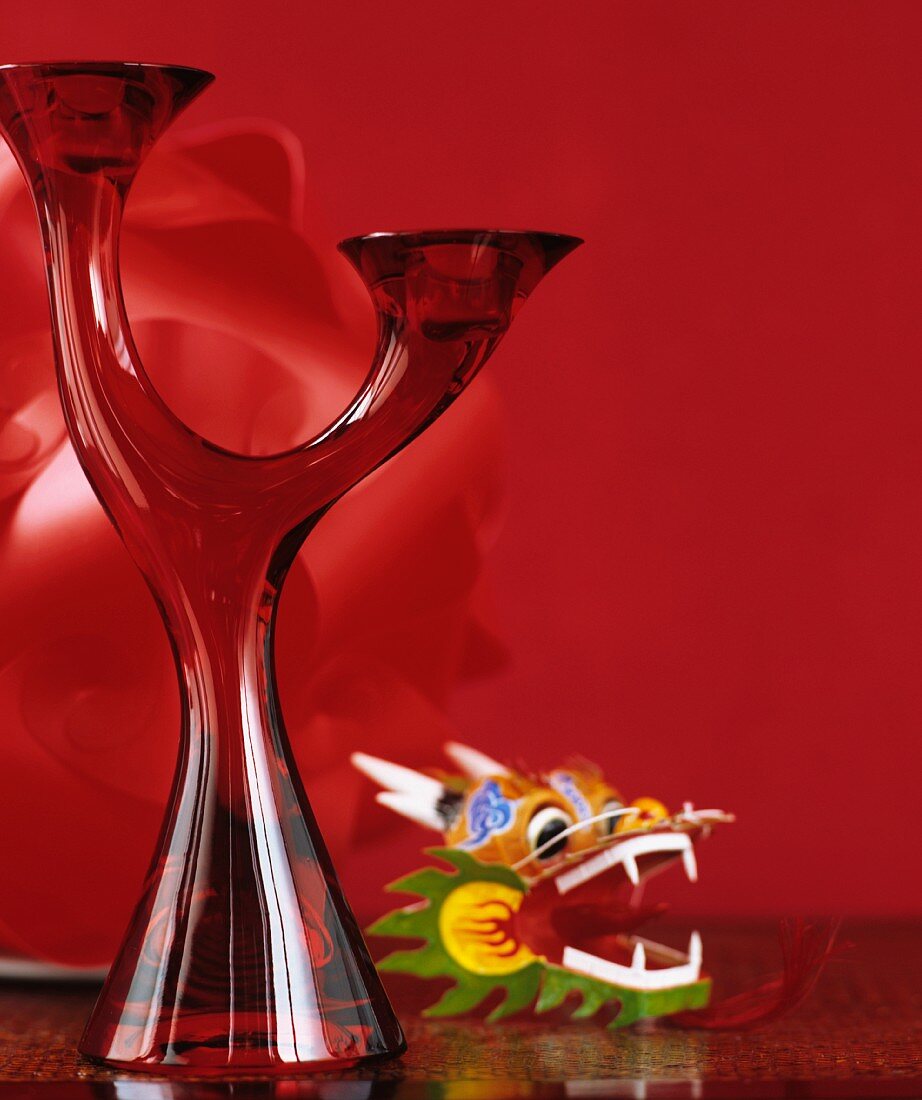 Zweiarmiger Kerzenständer aus rotem Glas neben Spielzeugfigur vor rotem Hintergrund