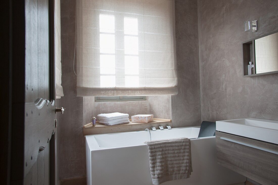 Designerbad - Blick durch offene Tür auf Badewanne vor Fenster mit heruntergezogenem Rollo und moderner Waschtischtrog an Wand