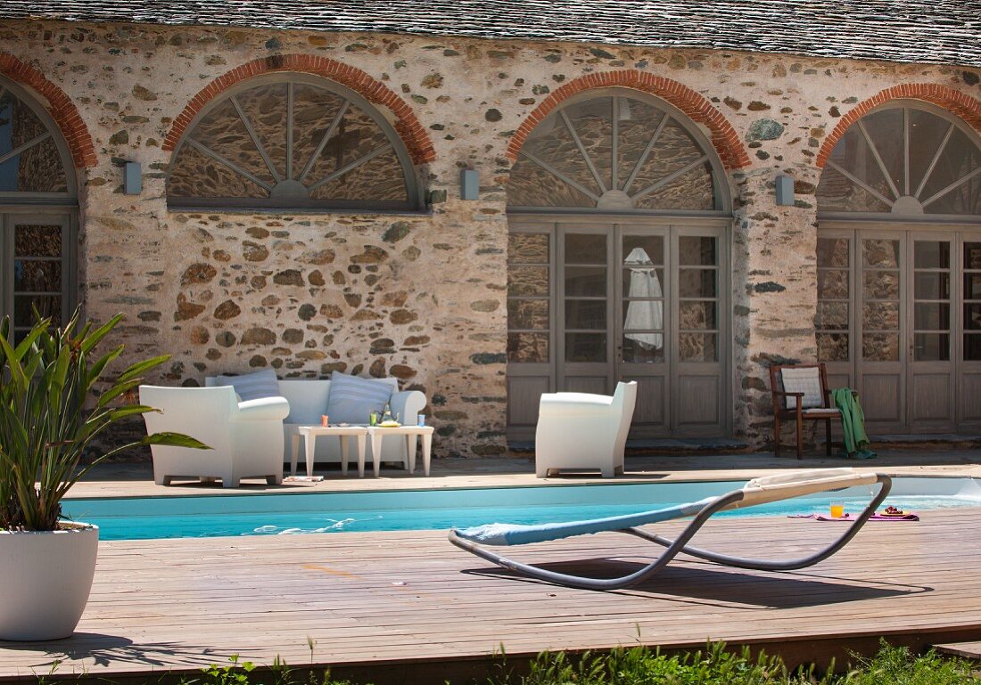 Designer lounger on wooden deck next to pool and white seating in front of Mediterranean house
