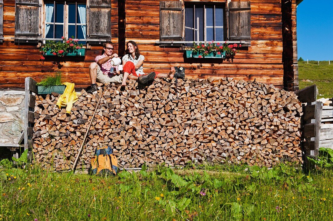 Couple sitting on stacked firewood in front of wooden house