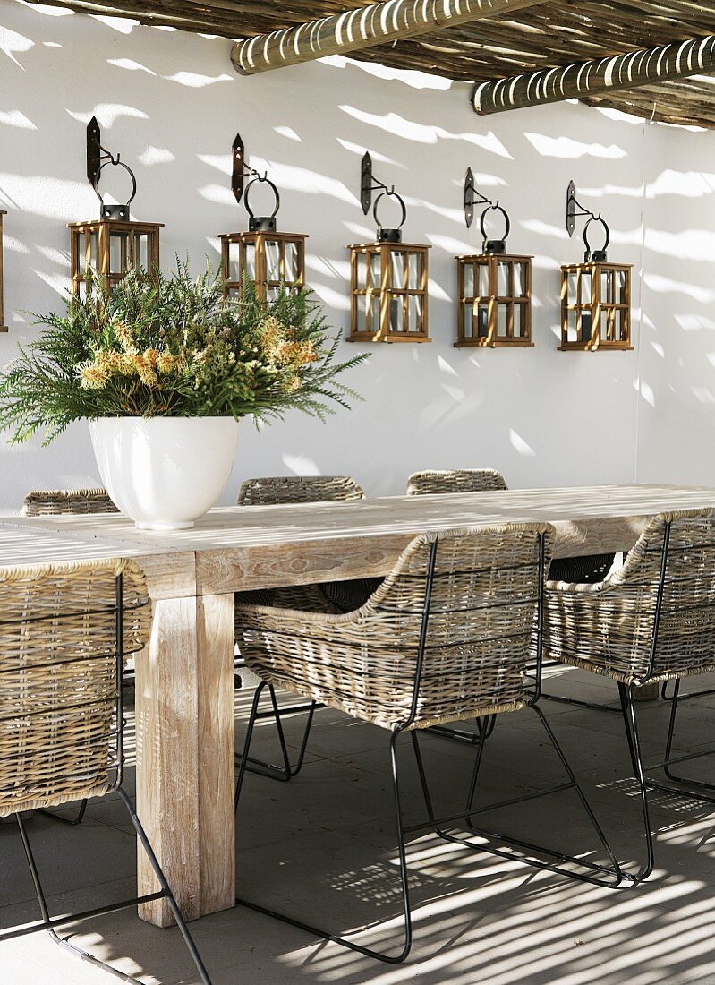 Lichteinfall durch Pergola auf Vintage Rattan Stühle mit Metallgestell an rustikalem Holztisch und Laternen an Hauswand aufgehängt
