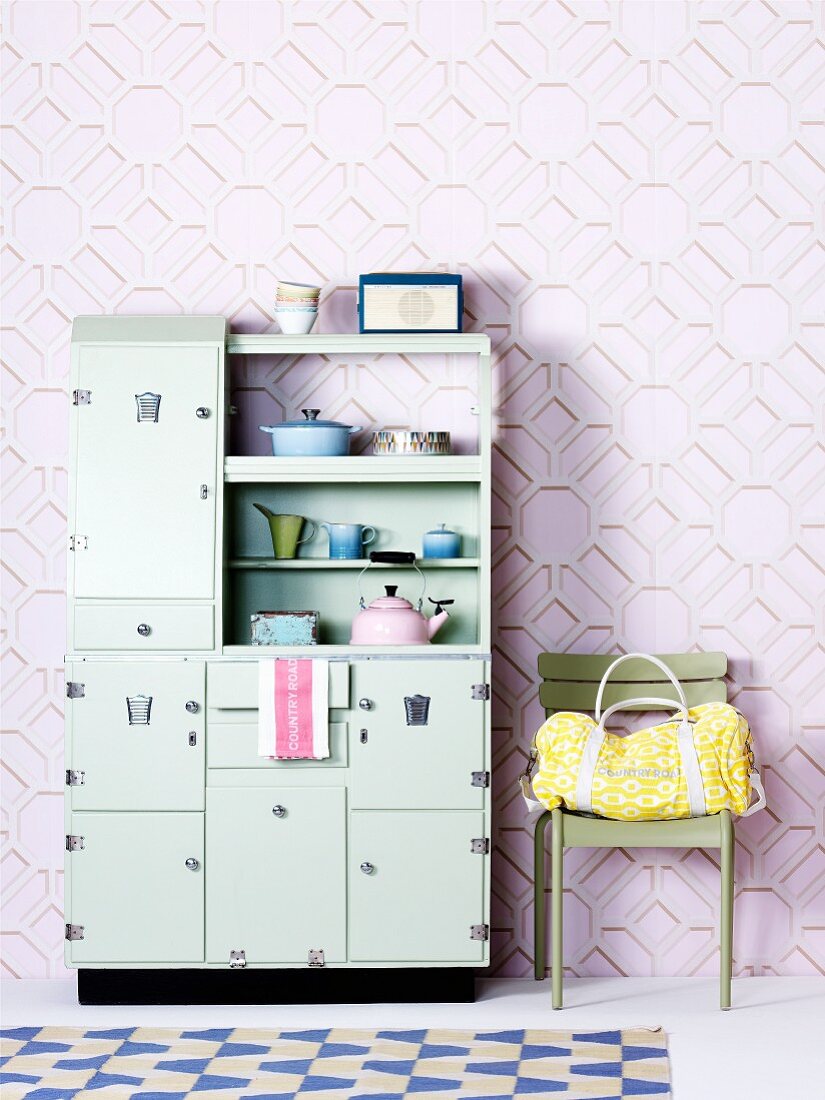 Fifties-style, pastel green kitchen dresser next to bag on chair against wallpaper with three-dimensional pattern