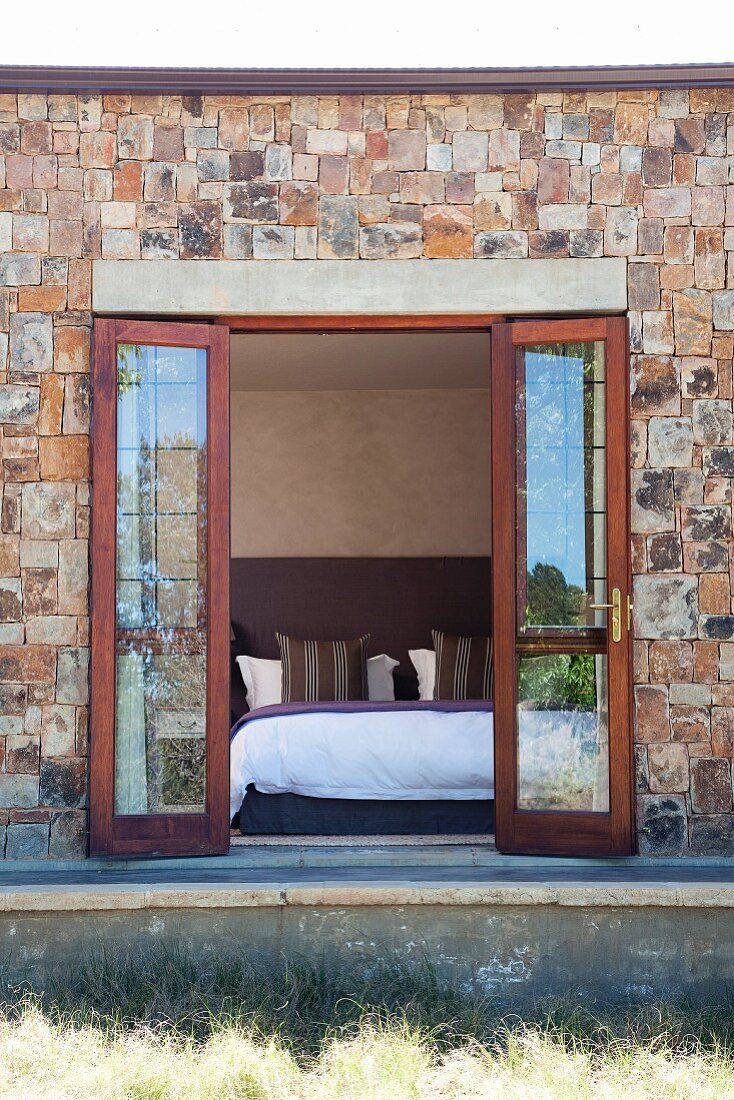 Open double terrace doors in stone facade with view into modern bedroom