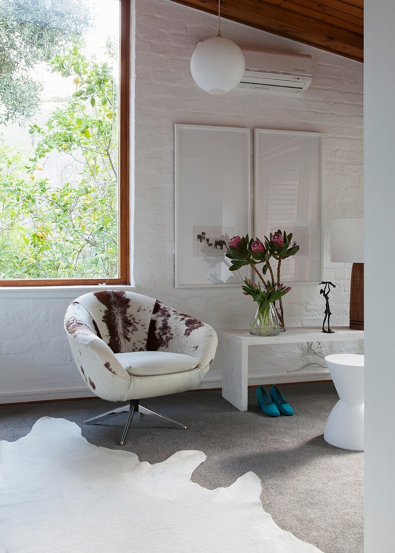 Swivel lounge chair with cow-skin cover next to window; framed pictures above white bench against whitewashed brick wall