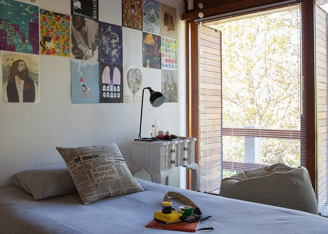 Einzelbett an Wand mit gepinnten Postern neben Vintage Nachttisch und geöffnete Jalousie an Balkontür