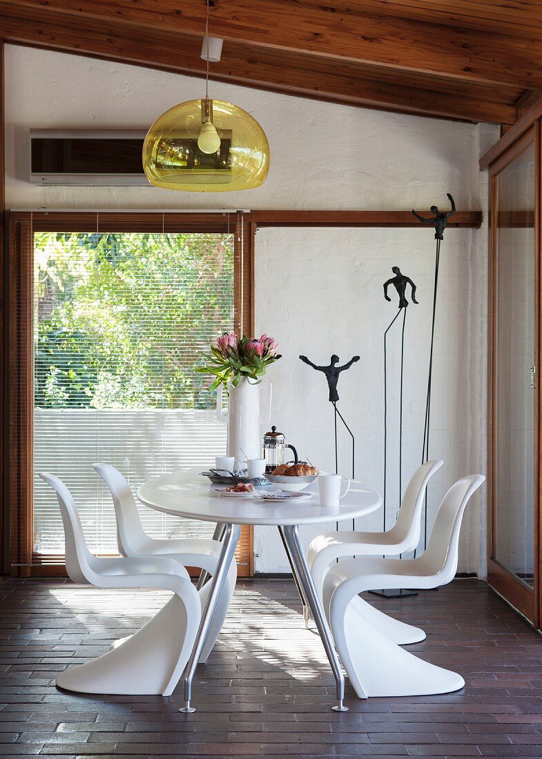 White, plastic shell chairs around table below pendant lamp with yellow, transparent lampshade in front of balcony door and metal sculptures