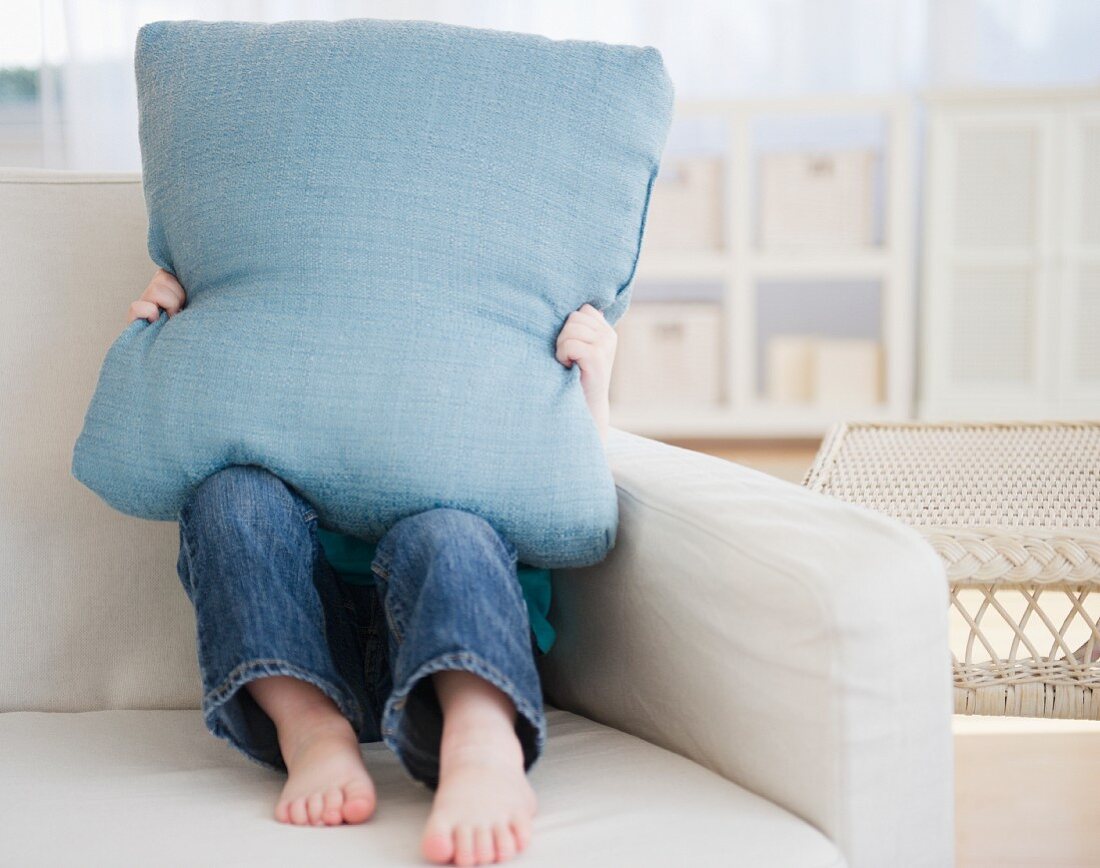Kleines Mädchen sitzt auf Sofa & versteckt sich hinter Kissen