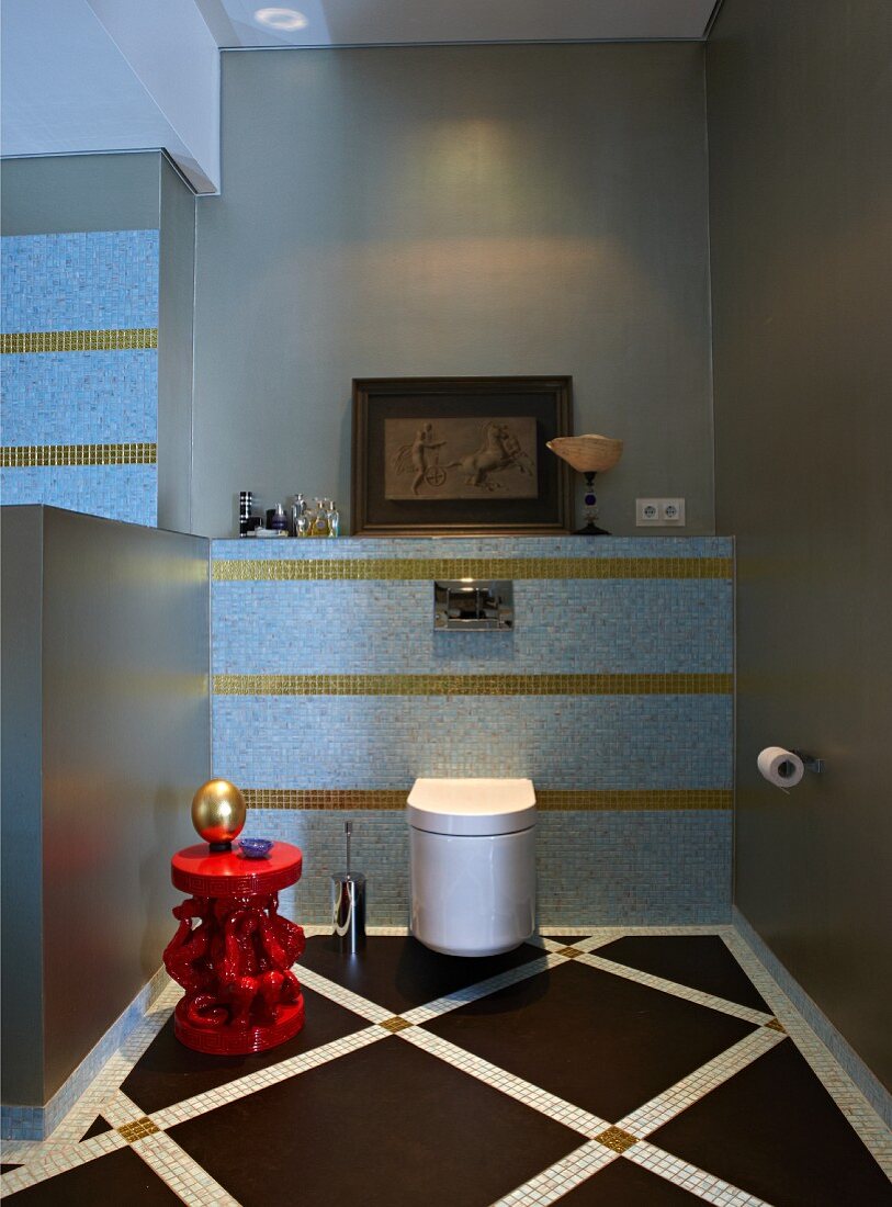 Wall-mounted toilet in elegant bathroom with pastel blue and gold mosaic tiles; gold ornament on red side table in foreground