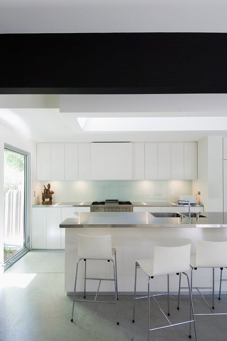 Bar stools with backs at a counter in a minimalist, white kitchen with large windows leading to the sunny garden
