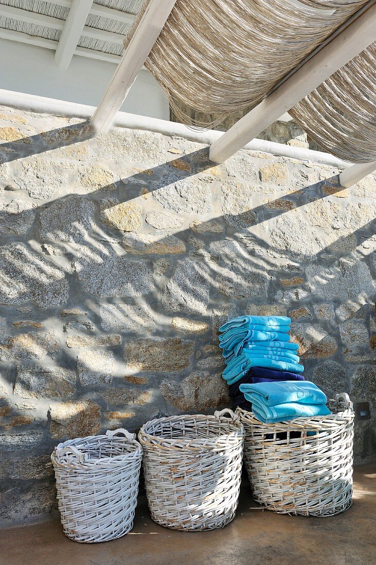 Stacked blue towels in one of three wicker baskets standing against stone wall on roofed terrace