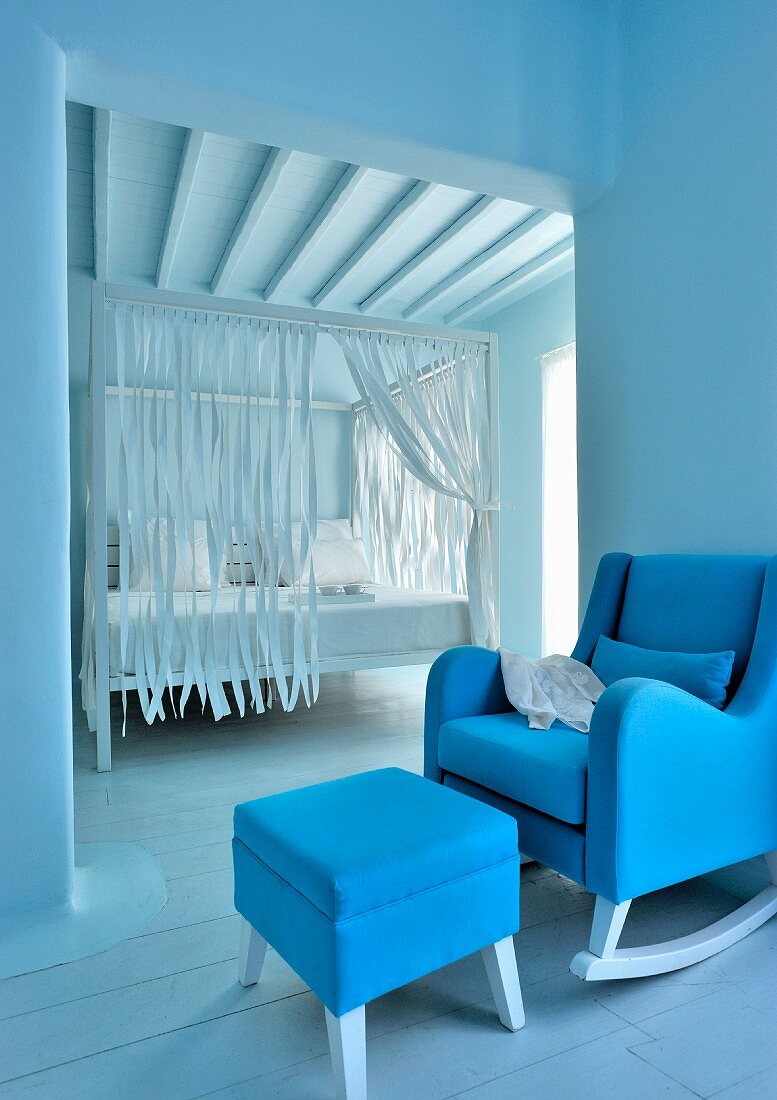 Gepolsterter Schaukelstuhl mit blauem Bezug und passendem Schemel vor Durchgang mit Blick auf weißem Himmelbett