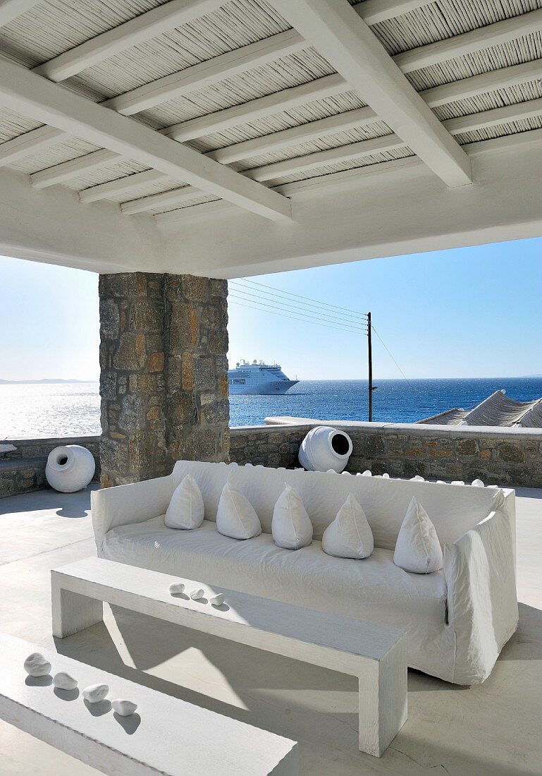 Drapierte Kissen auf Sofa mit weisser Husse und minimalistischer Tisch mit Bank auf überdachter Terrasse vor offenem Meer