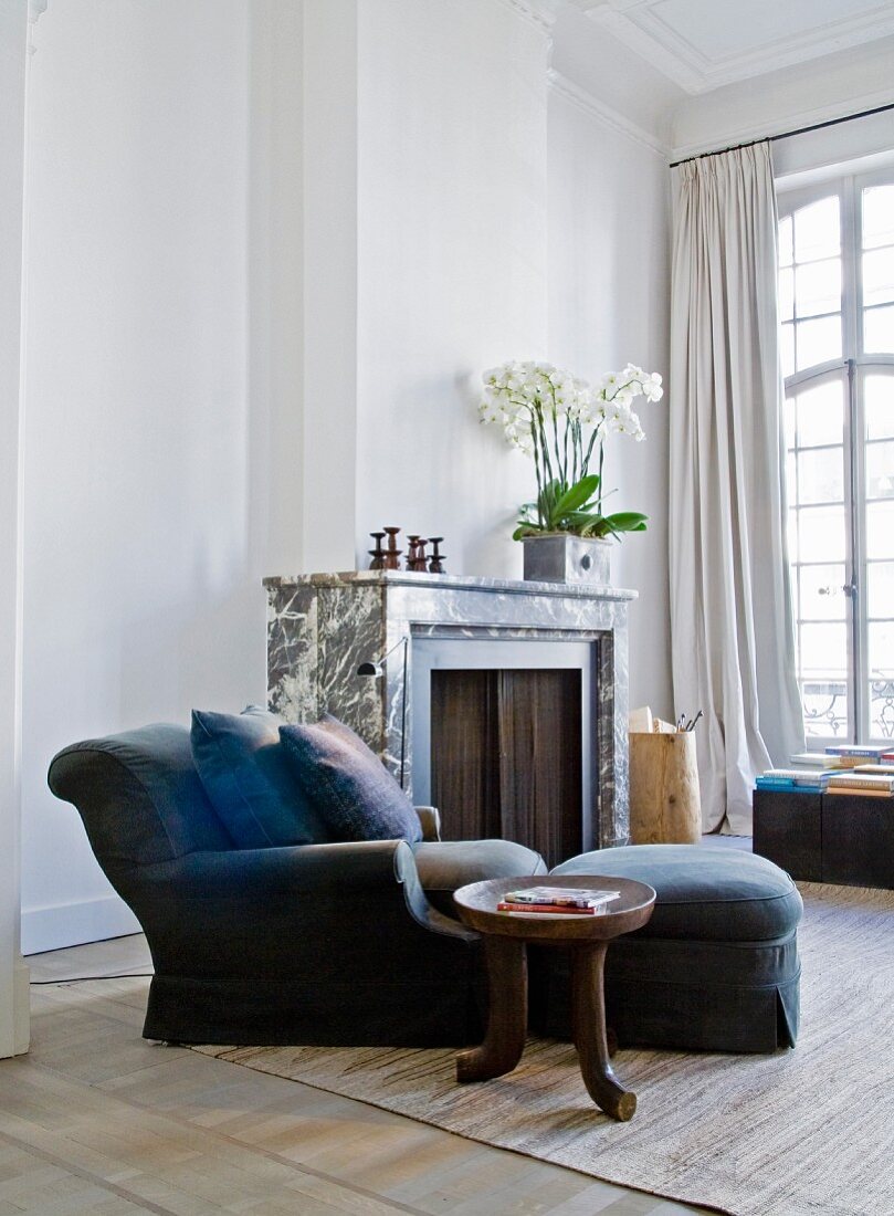 Beistelltisch im Ethnostil neben blauem Sessel mit passendem Fusshocker und Kissen vor Kamin in herrschaftlichem Wohnzimmer mit hohen Sprossenfenstern