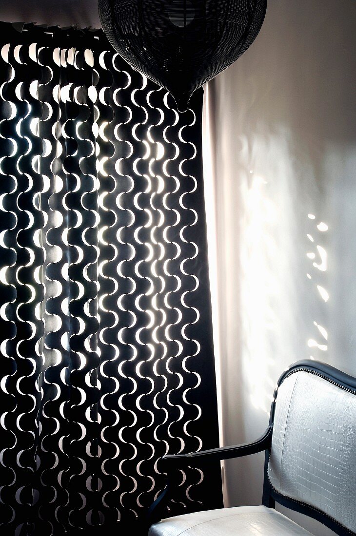 Dunkler Holzstuhl mit weisser Polsterung neben Vorhang mit weißem geschwungenem Muster auf dunklem Stoff