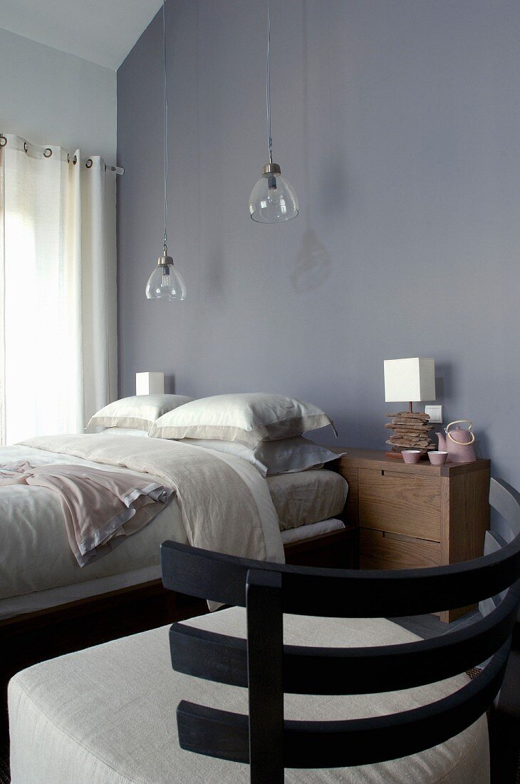 Sessel mit Holz Rückenlehne und naturfarbenem Polsterbezug vor Doppelbett unter Hängeleuchten mit Glasschirm vor grauer Wand