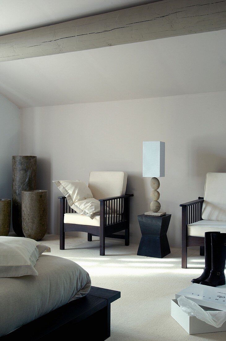 Table lamp on modern stool flanked by armchairs against grey well in bedroom with wood-beamed ceiling