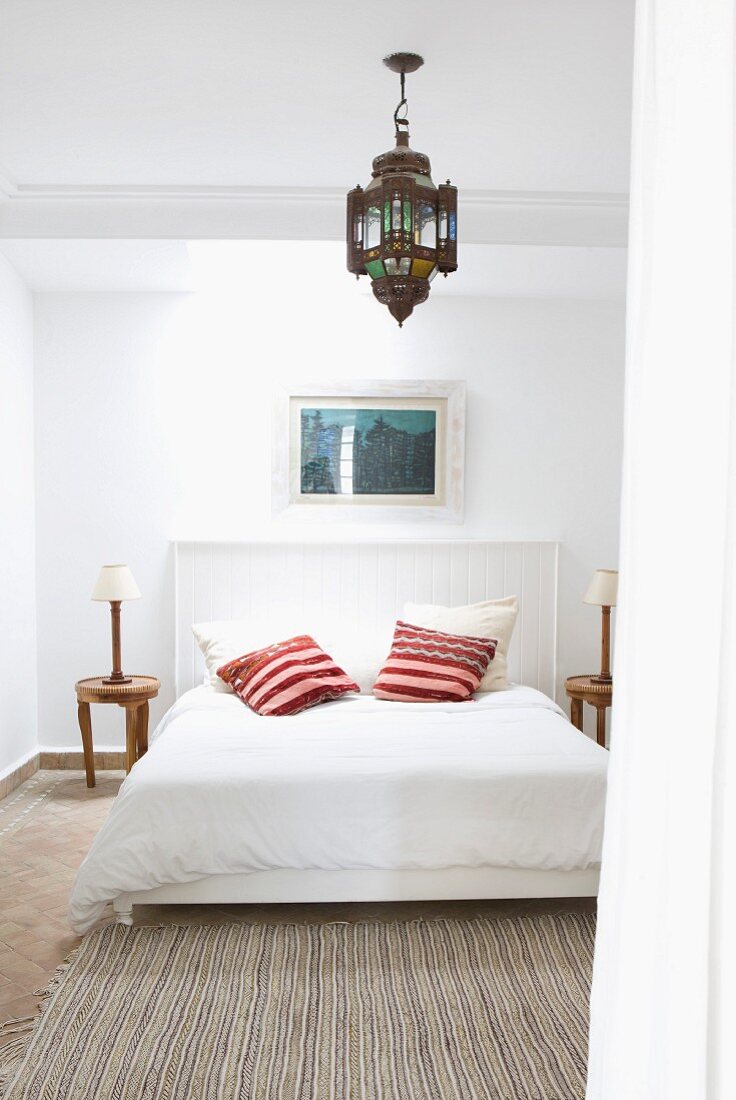 Simple bed with wood-panelled headboard and bright scatter cushions below Oriental pendant lamp