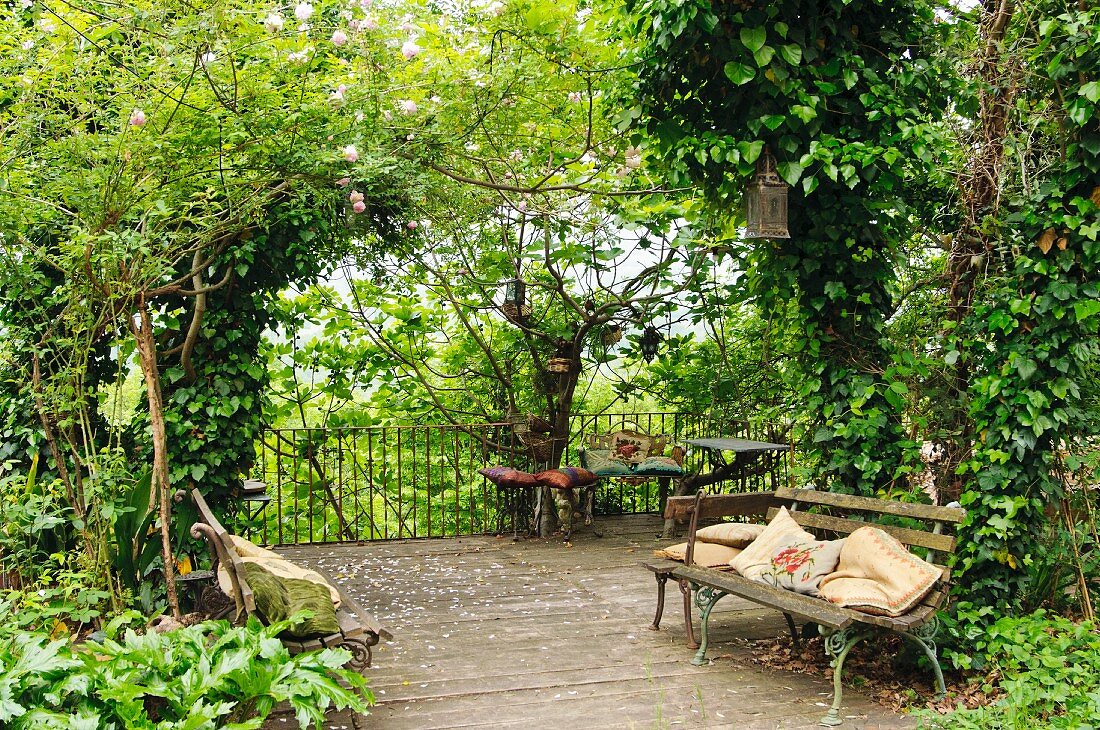 Vintage Gartenbank mit Kissen auf Holzterrasse in dicht bewachsenem Garten