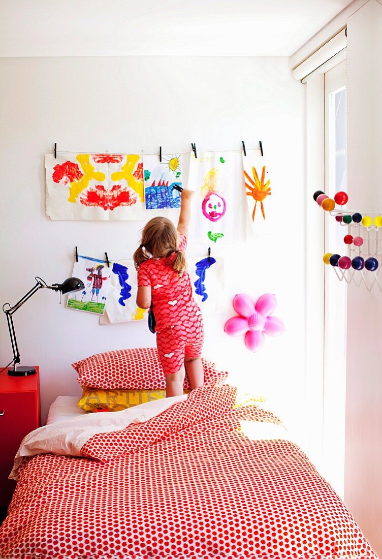 Mädchen steht auf Bett vor Wand mit aufgehängten, selbstgemalten Bildern