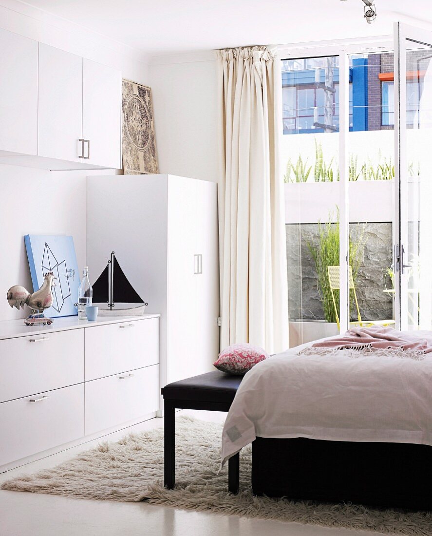 weiße Schränke und Bett mit Kleiderbank auf Flokati vor der offenen Balkontür eines Schlafzimmers