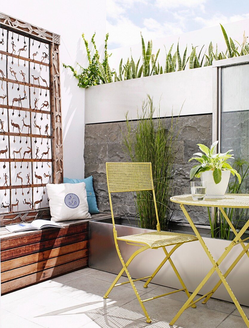 Artwork on wooden bench and wall with integrated planters behind sunny terrace with retro-style steel furniture