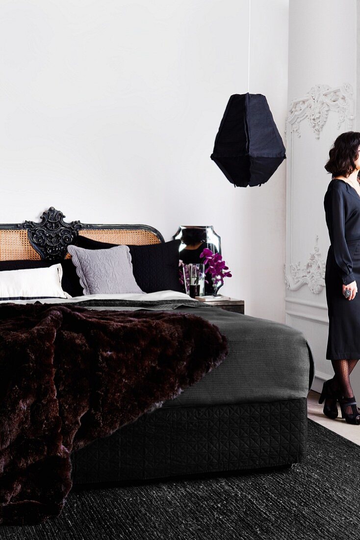 Schwarzes Bett mit geflochtenem, dekorativen Kopfende und einer luxuriösen Felldecke in elegantem Schlafzimmer mit anthrazitfarbenem Wollteppich und schwarzer Hängeleuchte