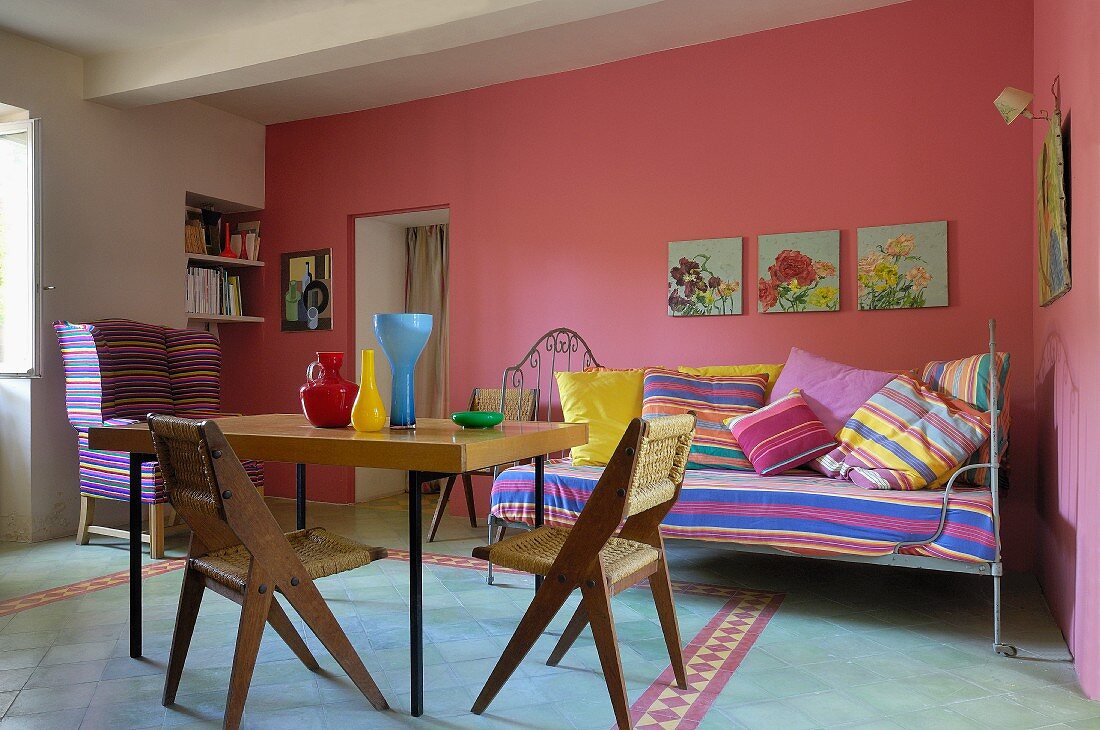 50s-style dining table and wooden chairs with wicker seats in front of daybed with striped cushions against pink-painted wall