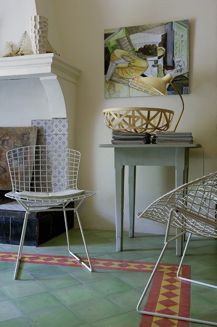 weiße Drahtgeflecht Stühle und rustikales Wandtischchen neben offenem Kamin im Wohnzimmer mit grünem Fliesenboden und Bordüre