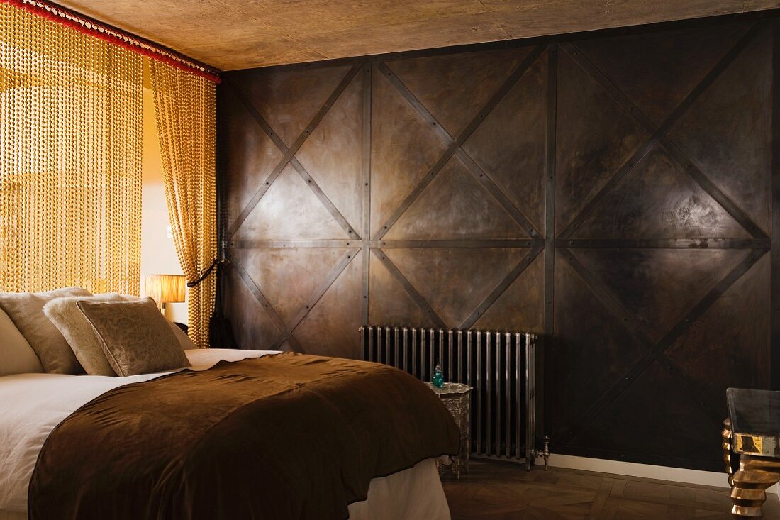 Doppelbett neben messingfarbenem Kettenvorhang und dunkler Holzpaneelwand in Herren Schlafzimmer
