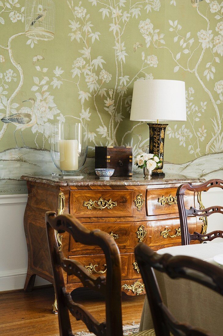 Barocke Kommode vor floraler Tapete und kunsthandwerklich bearbeitete Esszimmerstühle