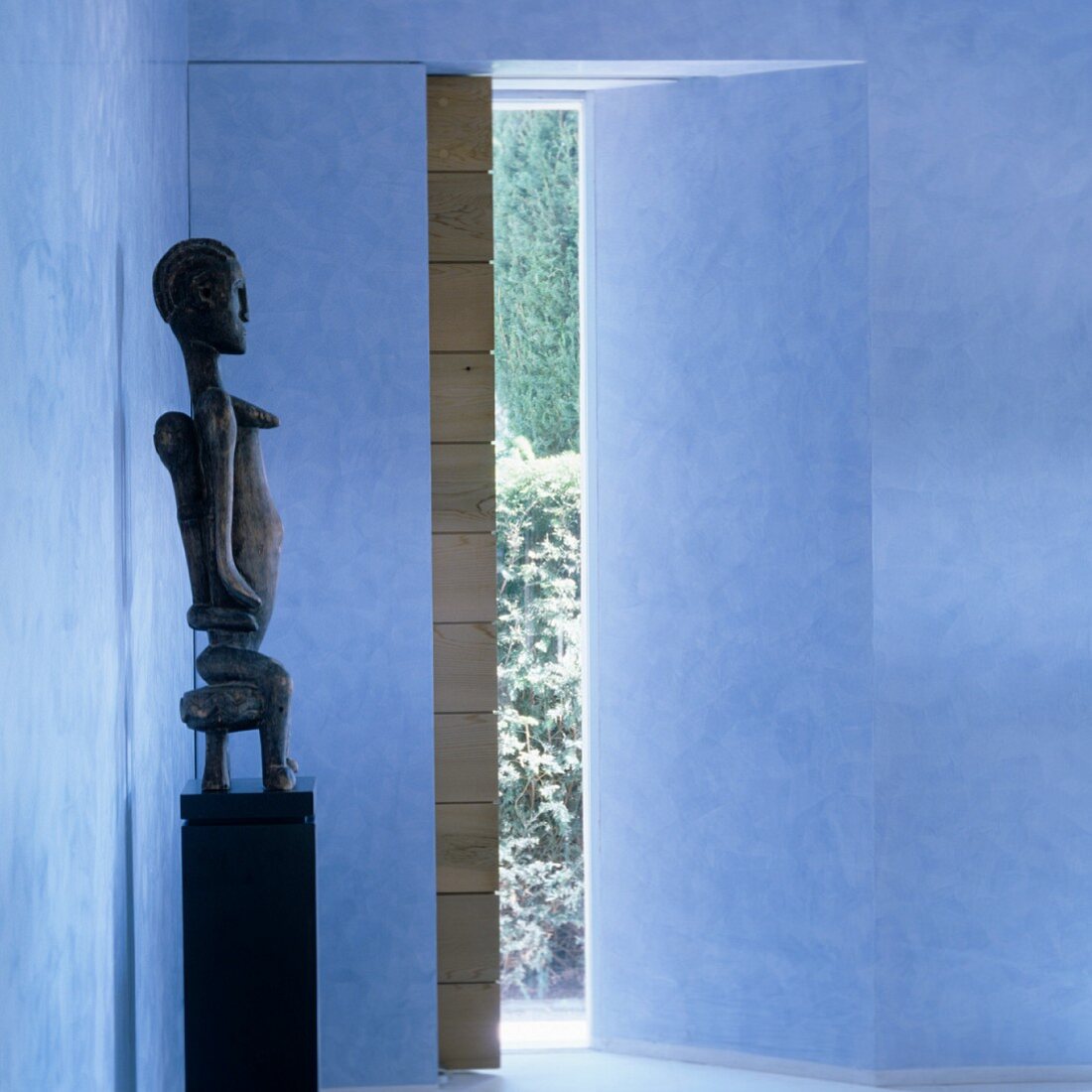 Afrikanische Skulptur auf Podest in blauem Vorraum mit Blick in Garten und Holzschiebetür