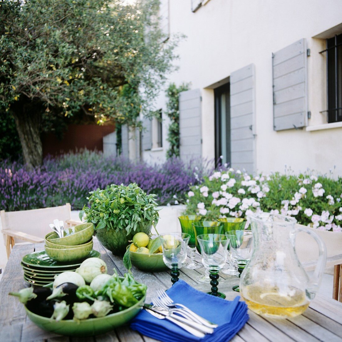 Aubergines, herbs and cutlery on wooden table outside modern Mediterranean holiday home; flowering lavender in background