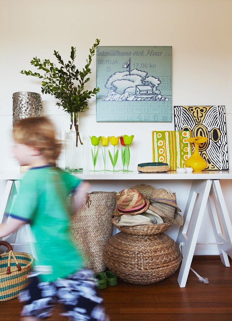 Laufendes Kind vor einfachem Wandtisch mit fröhlichen Bildern, Blumenvase und bunten Dekorationsgegenständen; unter dem Tisch verschiedene Körbe, Hüte und Kinderschuhe