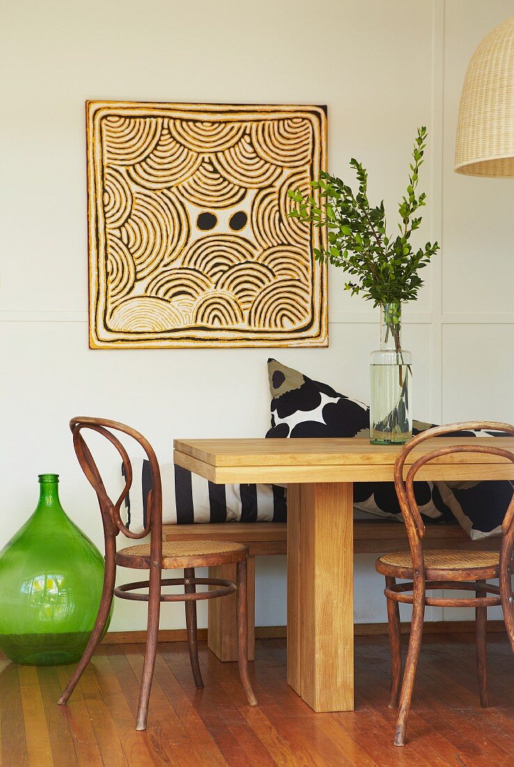 Expressives, quadratisches Wandbild; im Vordergrund ein massiver Holztisch mit Sitzbank und Vintage Bistrostühlen; eine alte Ölflasche dient als Bodenvase