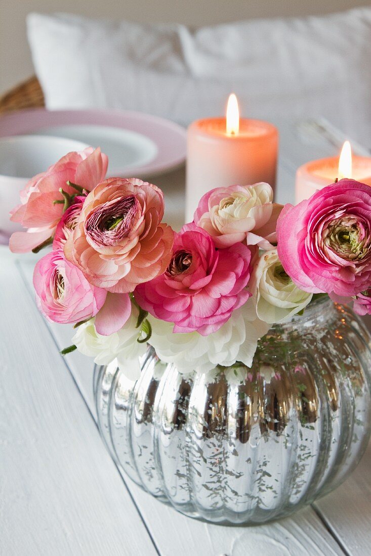 Ranunkelstrauss in silber glänzender Vase vor brennenden Kerzen