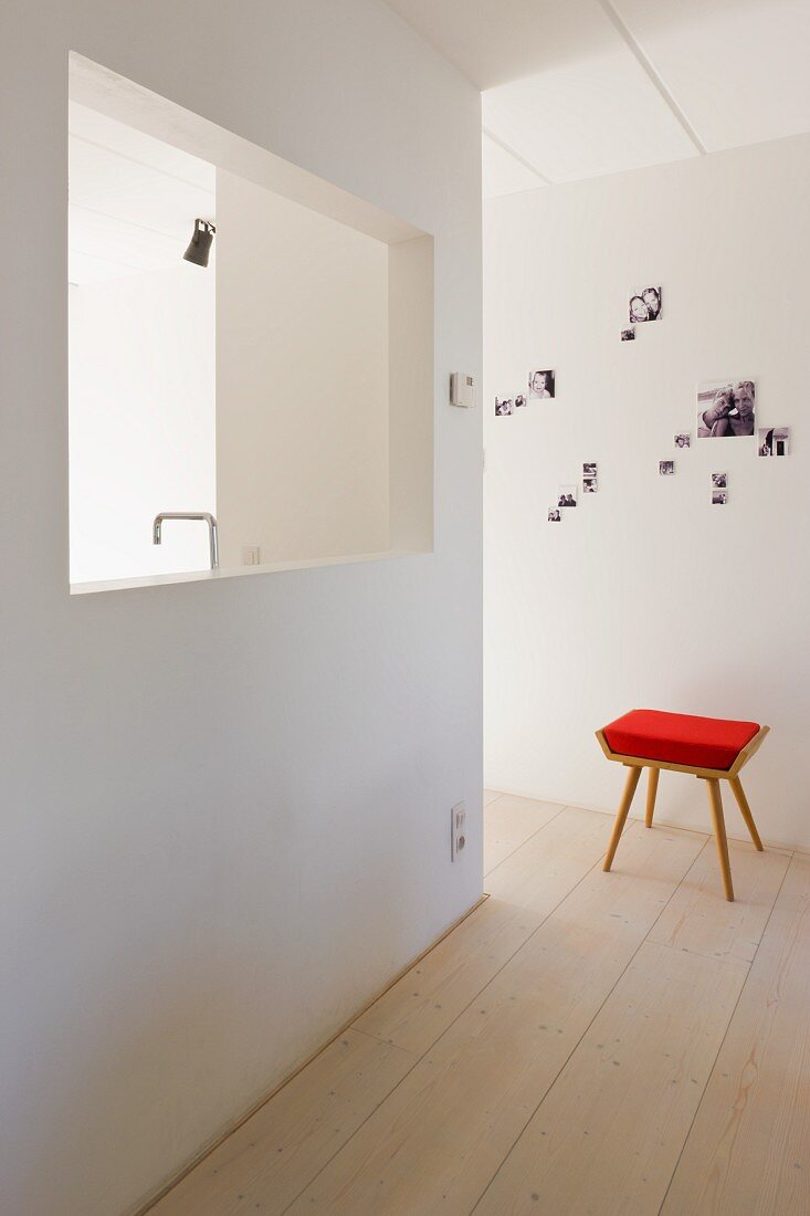 Hocker mit rotem Polster in schlichtem, minimalistischem Vorraum und fensterartige Öffnung in Wand mit Blick auf Küchenarmatur