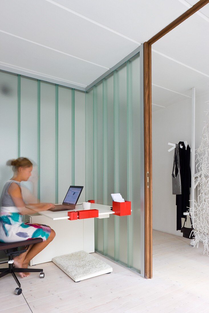 Frau an modernem Schreibttisch in offener Zimmerecke mit Glaswänden und Blick auf Garderobe im Gang