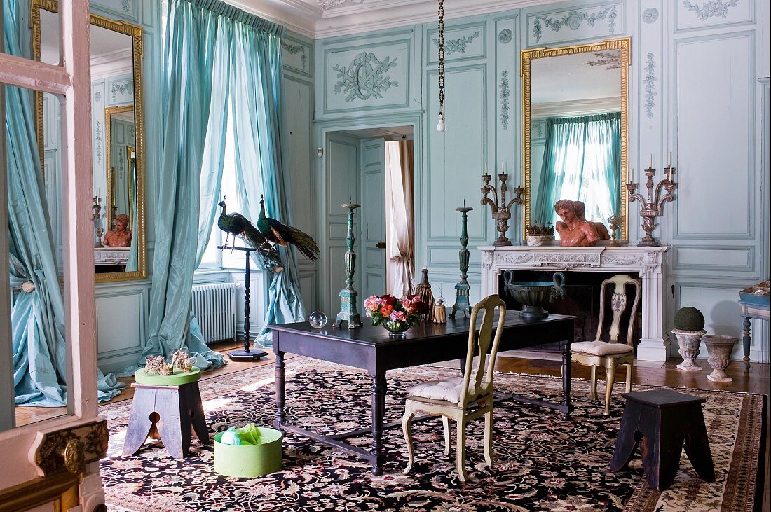 Herrschaftlicher Salon mit bemalter Holzpaneelwand und eingelassenem Spiegel über offenem Kamin, gegenüber Arbeitstisch mit Rokoko Stühlen auf gemustertem Teppich