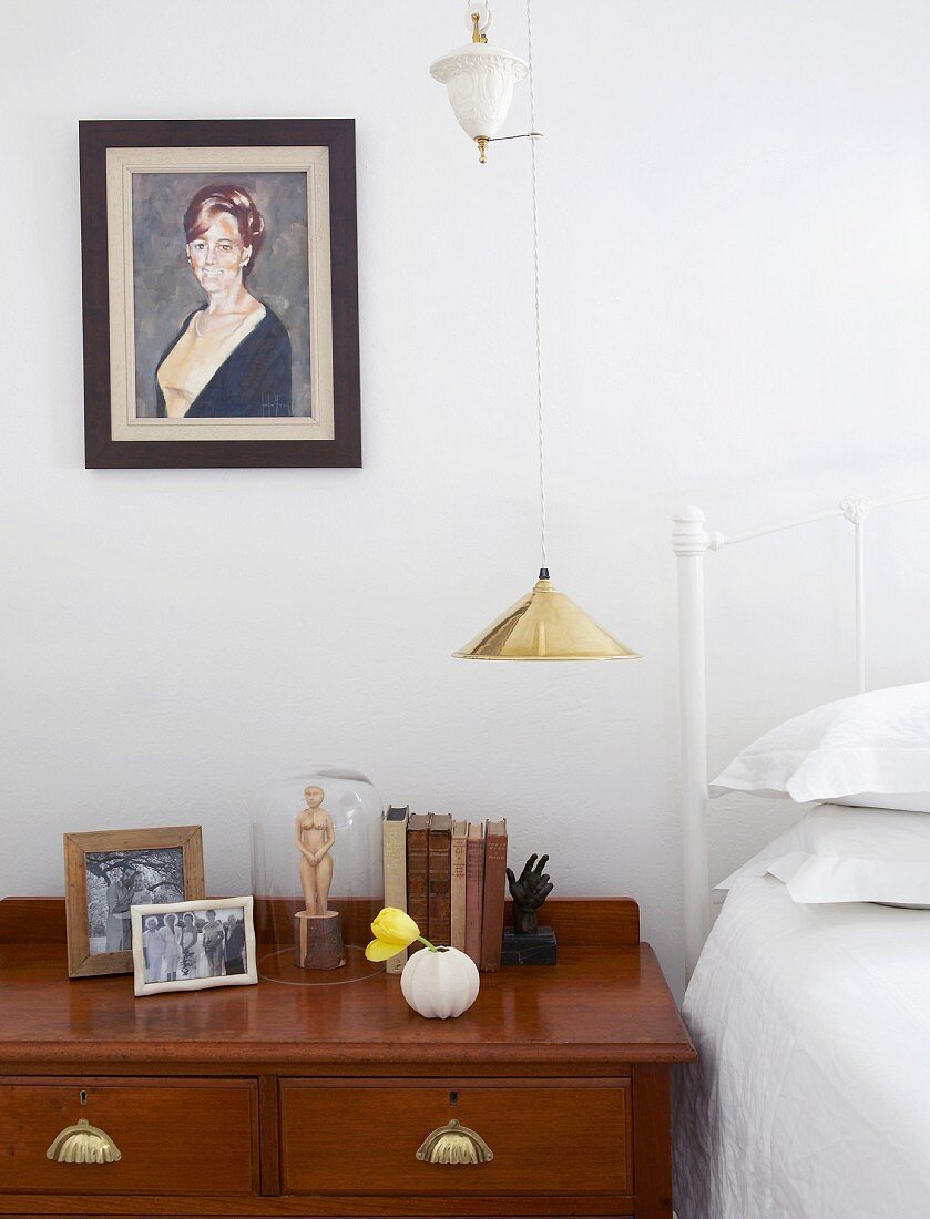Antique chest of drawers in bedroom with white metal bed; portrait of woman on wall next to gold pendant lamp