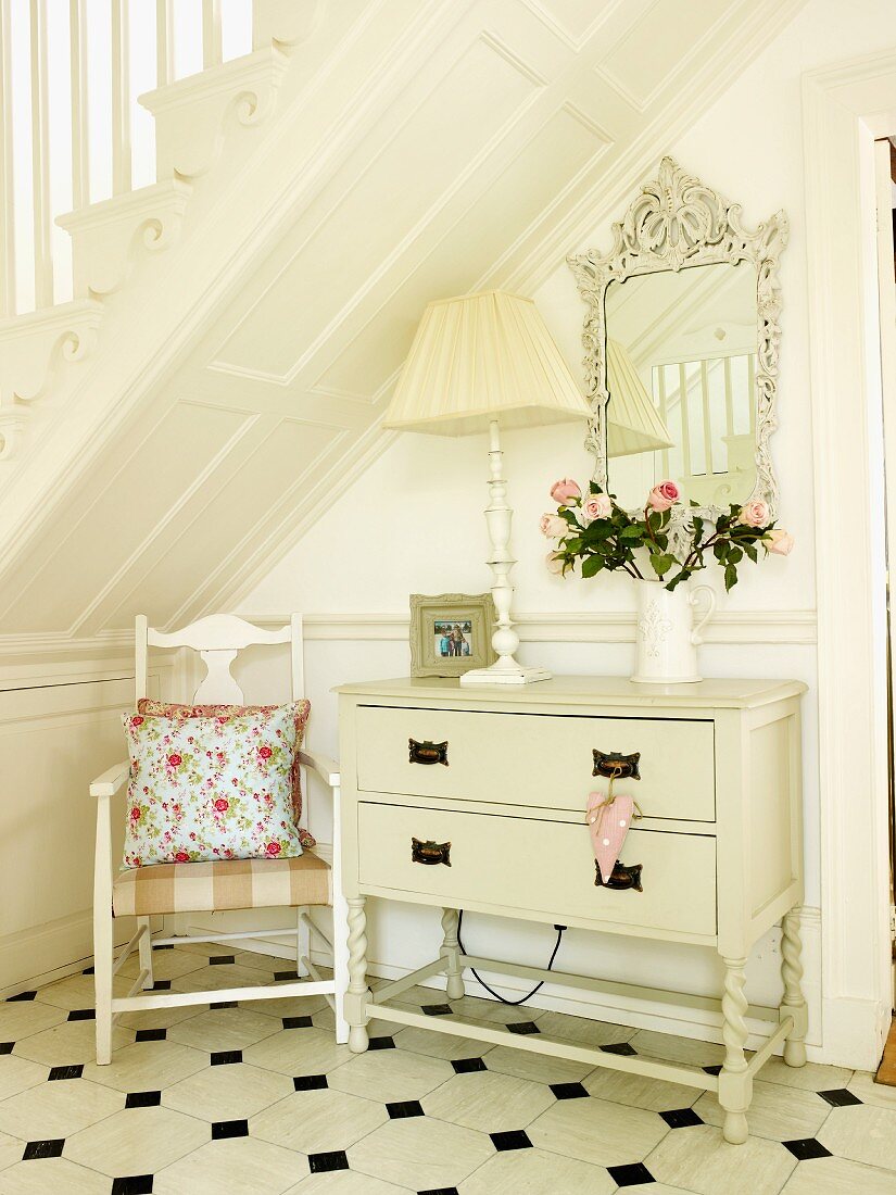 Stuhl mit Kissen und weiss lackierte Kommode mit Lampe, Blumenvase und Wandspiegel unter einer Holztreppe im Flur