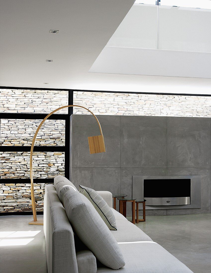 Pale grey sofa in front of wooden designer arc lamp in contemporary interior with open fireplace in exposed concrete wall