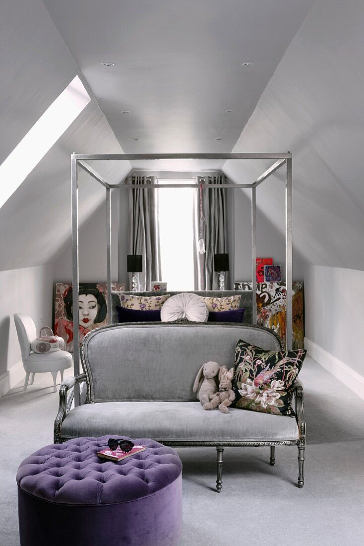 Metal four-poster bed, silver grey couch at foot of bed and purple ottoman in attic room with sloping ceiling