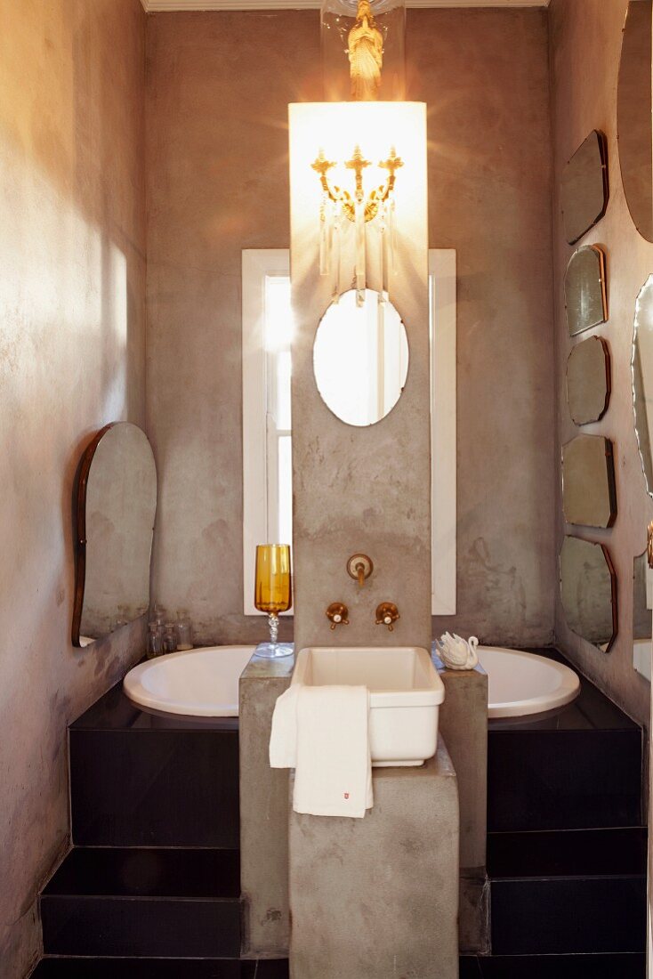 Freistehende Waschsäule aus Beton mit Wandkerzenleuchter über Spiegel vor schwarzem Podest mit eingelassener Badewanne